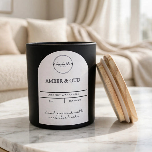 Amber & Oud luxe soy wax candle in a black jar with the wooden lid leaning beside it, styled on a marble coffee table in a bright living room. Clean non-toxic candle hand poured with 100% essential oils, Burbelle Boutique.