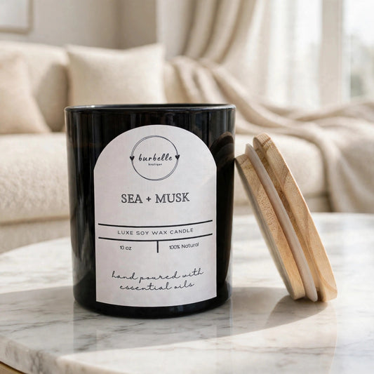 Sea + Musk luxe soy wax candle in a black jar with the wooden lid leaning beside it, styled on a marble coffee table in a bright living room. Clean non-toxic candle hand poured with 100% essential oils, Burbelle Boutique.
