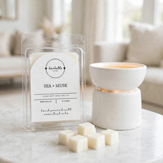 Sea + Musk luxe soy wax melts package beside a white ceramic wax warmer turned on with melted wax in the dish and loose cubes in front. Clean non-toxic wax melts hand poured with 100% essential oils, Burbelle Boutique.
