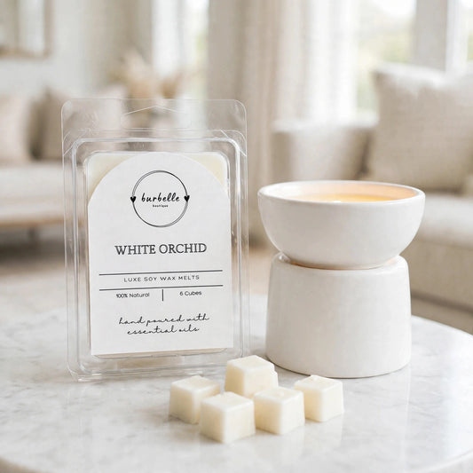 White Orchid luxe soy wax melts package beside a white ceramic wax warmer turned on with melted wax in the dish and loose cubes in front. Clean non-toxic wax melts hand poured with 100% essential oils, Burbelle Boutique.